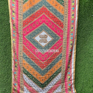 Peut inclure: Une textile aux motifs vifs et géométriques. Le motif présente un losange central avec des rayures alternées roses, oranges, vertes et rouges. Les bords sont ornés d'or et le texte "KHANTEXTILE7" est visible.