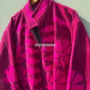 May include: A magenta velvet jacket with floral embroidery. The jacket has a mandarin collar and a button closure. The text "KHANTEXTILE7" is visible on the front. The jacket is hanging on a wooden hanger.