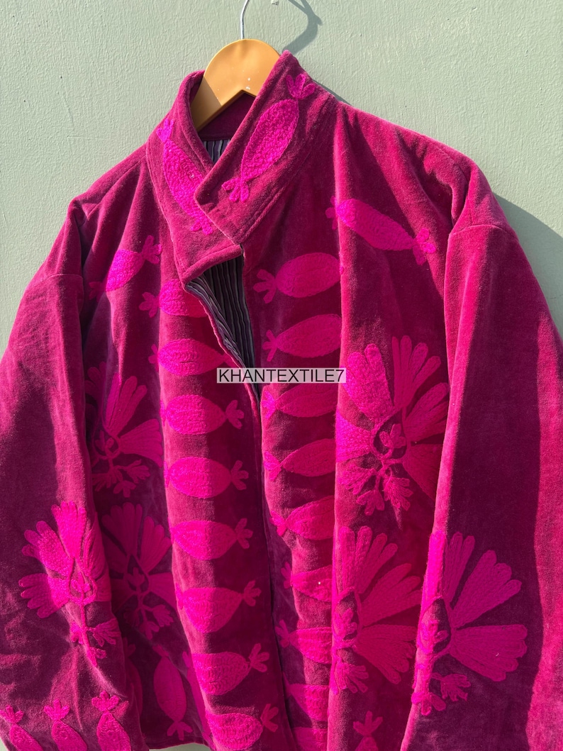 May include: A magenta velvet jacket with floral embroidery. The jacket has a mandarin collar and a button closure. The text "KHANTEXTILE7" is visible on the front. The jacket is hanging on a wooden hanger.