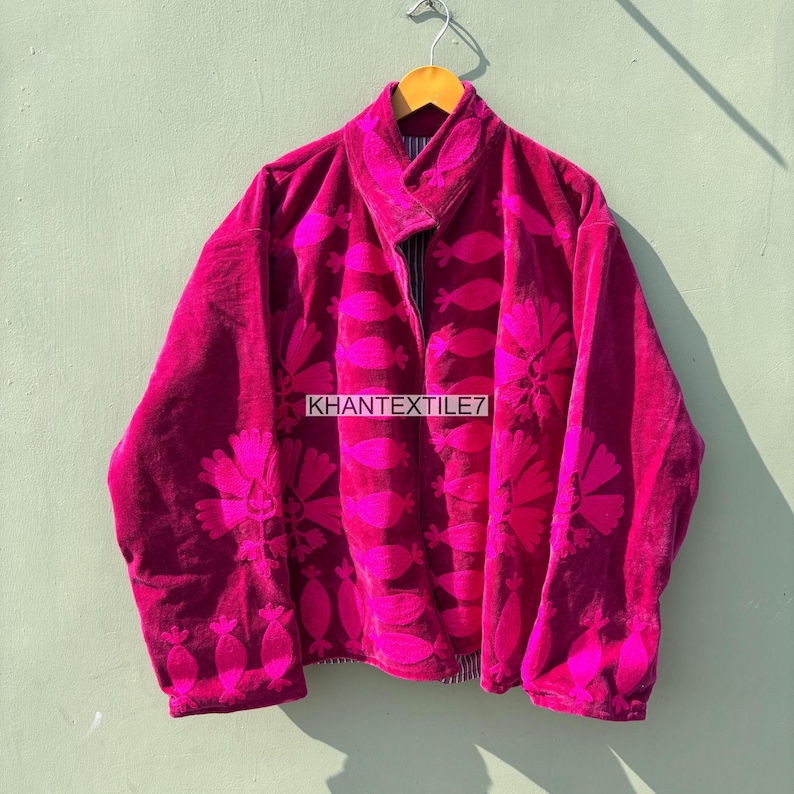 May include: A magenta velvet jacket with floral embroidery. The jacket has a wide collar and long sleeves. The text "KHANTEXTILE7" is visible on the front of the jacket. The jacket is hanging on a wooden hanger.