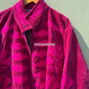 May include: A magenta velvet jacket with embroidered floral and leaf patterns. The jacket features a notched collar and a zipper closure. The text "KHANTEXTILE7" is visible on the jacket.