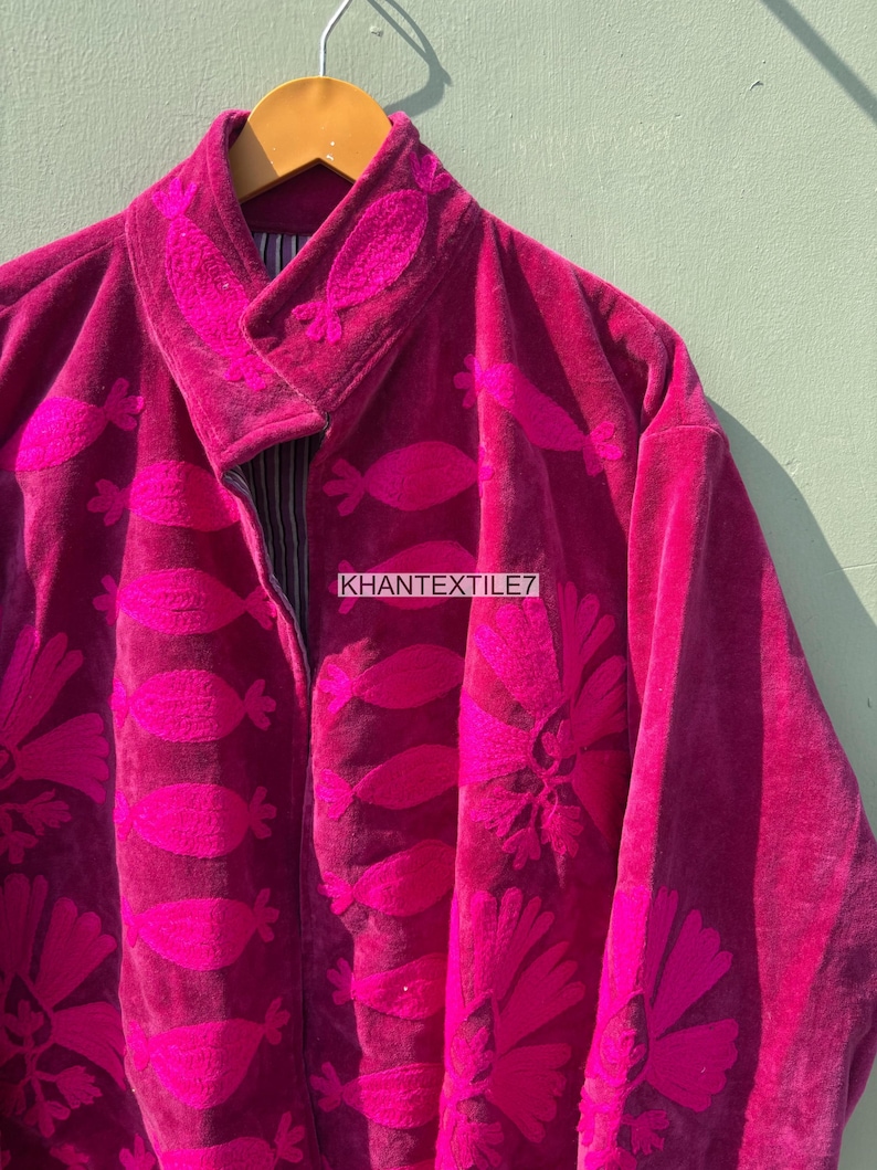 May include: A magenta velvet jacket with embroidered floral and leaf patterns. The jacket features a notched collar and a zipper closure. The text "KHANTEXTILE7" is visible on the jacket.