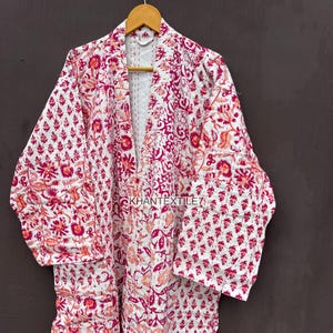 Puede incluir: Bata con estampado floral blanco y rosa colgada de una percha de madera. La bata presenta un diseño acolchado con cuello en V y mangas largas. El texto "KHANTEXTILE7" es visible en la bata.