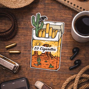 Puede incluir: Una pegatina de un paquete de cigarrillos con un dise&ntilde;o de paisaje des&eacute;rtico, con un cactus y un cuervo. Se muestra el texto "20 Cigarettes". La imagen est&aacute; rodeada por un sombrero de vaquero, gafas de sol y otros accesorios.