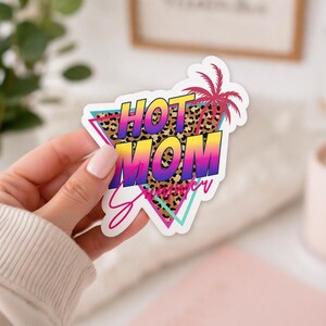 以下が含まれることがあります： レトロなデザインの「HOT MOM Summer」という言葉が入ったカラフルなステッカー。テキストは黄色、オレンジ、紫で、ヒョウ柄の背景があります。ピンクのヤシの木とターコイズのアクセントがデザインを完成させています。