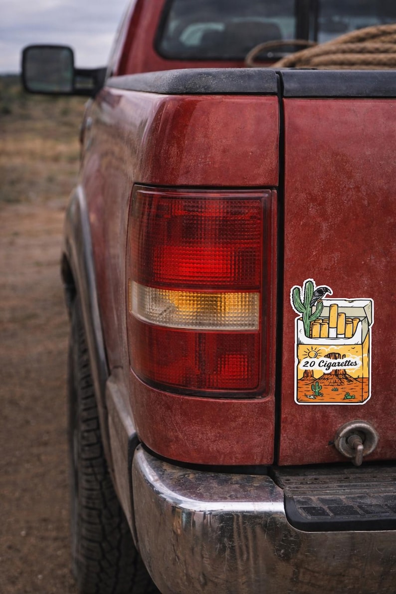 Puede incluir: Primer plano de la parte trasera de una camioneta roja con una pegatina de un paquete de cigarrillos con un paisaje des&eacute;rtico y el texto "20 Cigarettes". La luz trasera de la camioneta es visible.