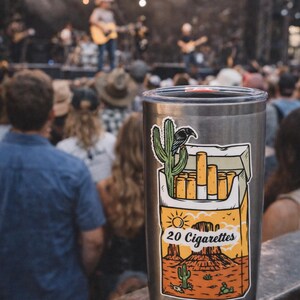 Puede incluir: Un vaso de acero inoxidable con una pegatina que representa un paisaje des&eacute;rtico, un cactus, un cuervo y un paquete de cigarrillos. El texto "20 Cigarettes" es visible. El fondo muestra un concierto.