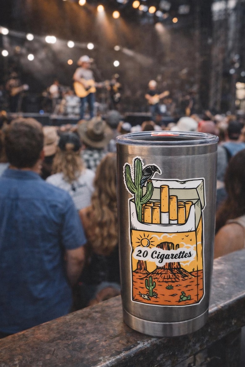 Puede incluir: Un vaso de acero inoxidable con una pegatina que representa un paisaje des&eacute;rtico, un cactus, un cuervo y un paquete de cigarrillos. El texto "20 Cigarettes" es visible. El fondo muestra un concierto.