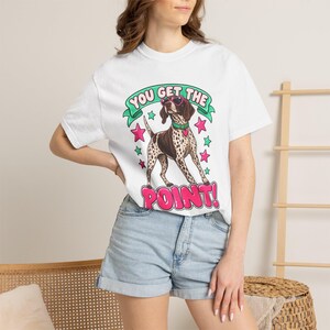 May include: A white short-sleeved t-shirt featuring a graphic of a brown and white spotted dog wearing pink heart-shaped sunglasses and a green collar. The text "YOU GET THE POINT!" is printed in pink and green with pink and green star accents.