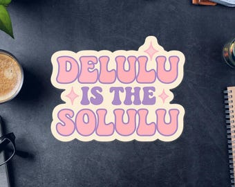 May include: A sticker with the text "DELULU IS THE SOLULU" in a retro font. The text is in pink and purple, with a cream-colored border. The sticker is on a dark surface with other objects.