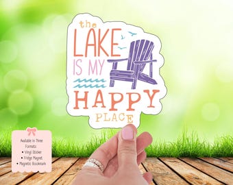 May include: A sticker with the text "The Lake Is My Happy Place" in coral and purple. The design includes a purple Adirondack chair, waves, and birds. The sticker is held by a hand with a pink nail polish.