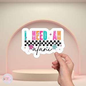 Puede incluir: Una pegatina blanca con el texto "I NEED AN afari" en letras coloridas y desgastadas. El dise&ntilde;o incluye un patr&oacute;n de tablero de ajedrez en blanco y negro, flores y estrellas. La pegatina est&aacute; sujeta por una mano.