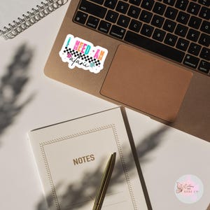 Puede incluir: Una pegatina colorida con el texto "I NEED AN alani", con un dise&ntilde;o de cuadros y detalles florales. Se encuentra sobre un ordenador port&aacute;til, con un cuaderno y un bol&iacute;grafo cerca.