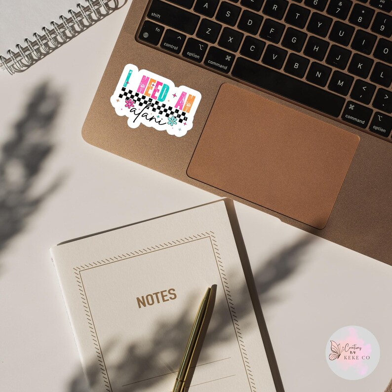 Puede incluir: Una pegatina colorida con el texto "I NEED AN alani", con un dise&ntilde;o de cuadros y detalles florales. Se encuentra sobre un ordenador port&aacute;til, con un cuaderno y un bol&iacute;grafo cerca.