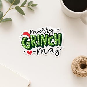May include: A sticker with the words "merry GRINCH mas" in green and white, with a red heart and a Santa hat on the letter "G". The sticker is on a white surface, with a cup of coffee and a ball of twine in the background.