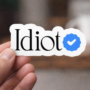 以下が含まれることがあります： 黒い文字で「Idiot」と書かれた白いステッカー。青いチェックマークのバッジが付いています。ぼやけた背景にステッカーが掲げられています。