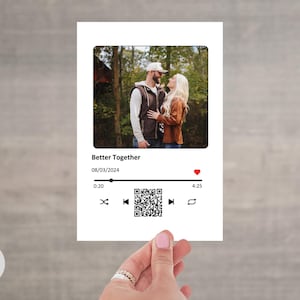 Puede incluir: Una tarjeta blanca con una foto de una pareja, el texto "Better Together" y una fecha. Debajo de la foto hay controles de reproductor de música, un código QR y un icono de corazón. La tarjeta está sostenida por una mano.