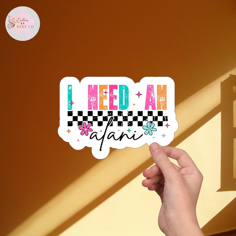 Puede incluir: Una pegatina blanca con el texto "I NEED AN safari" en letras coloridas de estilo retro. El dise&ntilde;o incluye un patr&oacute;n de cuadros blancos y negros, flores y estrellas. La pegatina est&aacute; sujeta por una mano sobre un fondo marr&oacute;n.