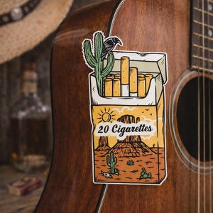 Puede incluir: Una pegatina de un paquete de cigarrillos con un dise&ntilde;o de paisaje des&eacute;rtico, con cactus y un cuervo, est&aacute; adherida a una guitarra ac&uacute;stica marr&oacute;n. El paquete dice "20 Cigarrillos". Un sombrero de vaquero es visible en el fondo.