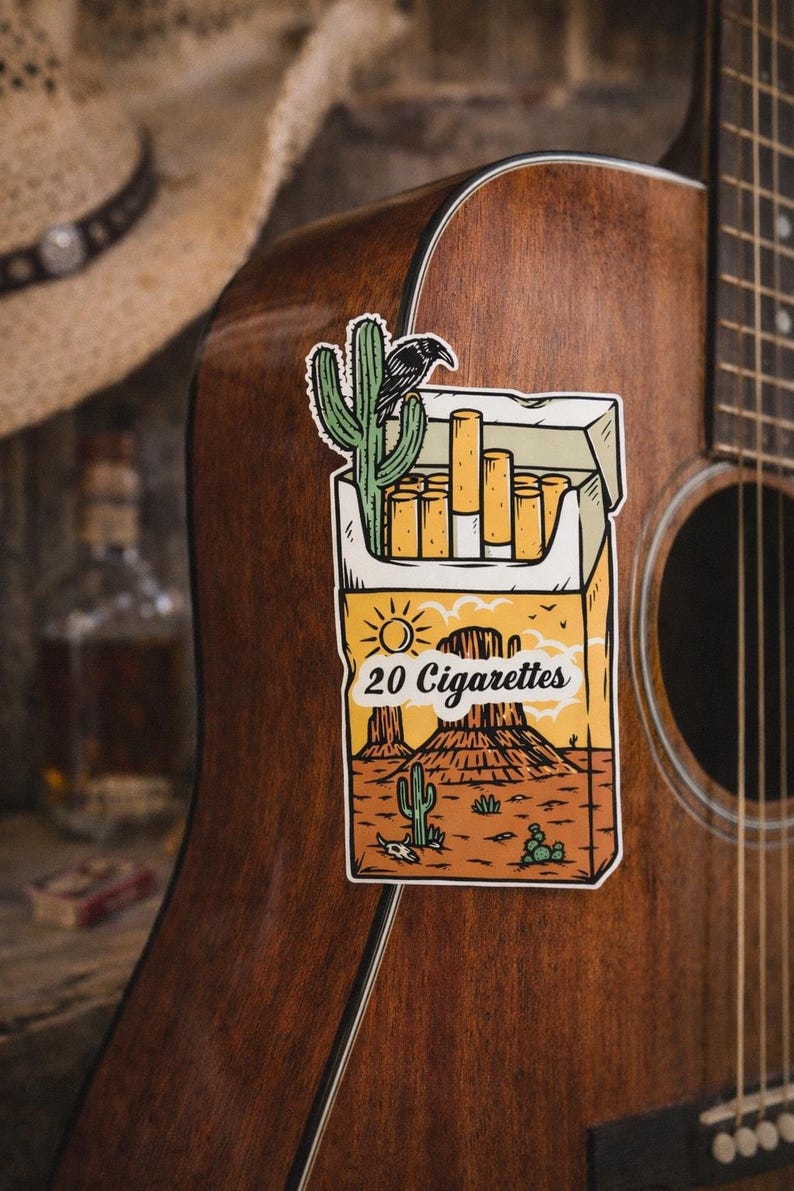 Puede incluir: Una pegatina de un paquete de cigarrillos con un dise&ntilde;o de paisaje des&eacute;rtico, con cactus y un cuervo, est&aacute; adherida a una guitarra ac&uacute;stica marr&oacute;n. El paquete dice "20 Cigarrillos". Un sombrero de vaquero es visible en el fondo.