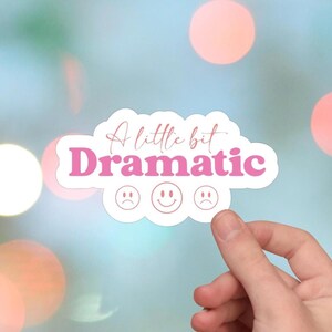 Może przedstawiać: Biała naklejka z napisem "A little bit Dramatic" w różowym skrypcie i pogrubioną czcionką. Poniżej tekstu znajdują się trzy emotikony: smutna buzia, uśmiechnięta buzia i kolejna smutna buzia.