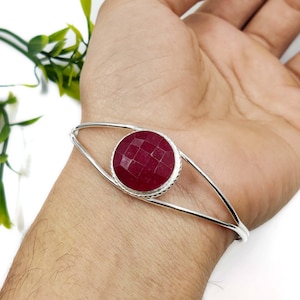 Pode incluir: Uma pulseira de prata com uma pedra preciosa circular facetada de cor vermelho-profundo no centro. A banda de metal divide-se em dois braços finos e lisos que se curvam à volta do pulso, encontrando-se na pedra engastada com facetas.