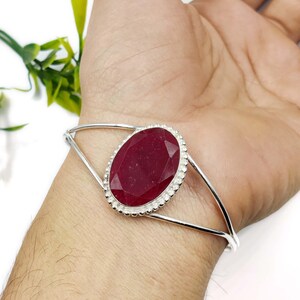 May include: A silver cuff bracelet featuring a large, oval-shaped, faceted deep red gemstone at the center. The stone is set in a decorative, beaded silver bezel, and the band consists of two thin, polished silver wires that meet at the setting.