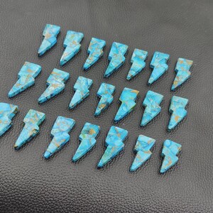 May include: A collection of turquoise-colored gemstone lightning bolt pendants. Each bolt features a unique pattern of blue and gold veining. The pendants are arranged on a textured black surface.