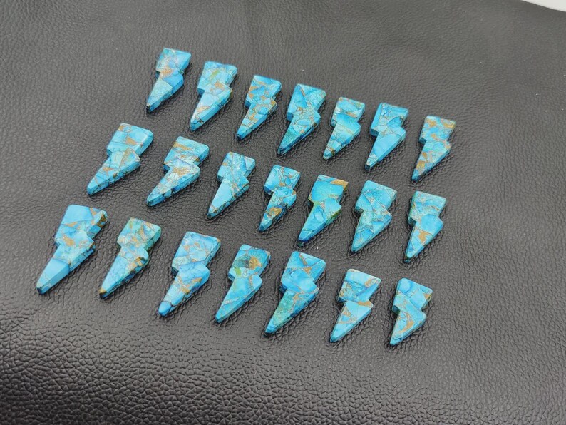 May include: A collection of turquoise-colored gemstone lightning bolt pendants. Each bolt features a unique pattern of blue and gold veining. The pendants are arranged on a textured black surface.