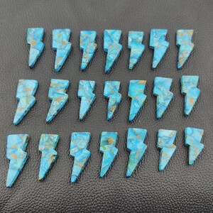 May include: Twenty-one turquoise-colored gemstone lightning bolt pendants with gold-colored veining. The pendants are arranged in three rows on a black textured surface. Each bolt is approximately 1 inch in length.