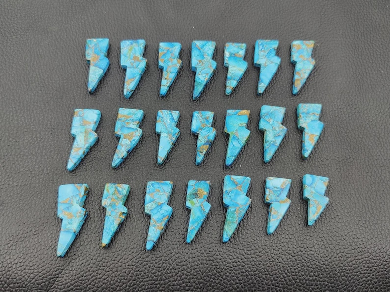 May include: Twenty-one turquoise-colored gemstone lightning bolt pendants with gold-colored veining. The pendants are arranged in three rows on a black textured surface. Each bolt is approximately 1 inch in length.