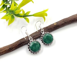 Op de afbeelding: Een paar zilveren oorbellen met een ronde, gefacetteerde, smaragdgroene edelsteen. De oorbellen hebben een decoratieve rand en een haaksluiting. De oorbellen worden gepresenteerd op een bruine tak tegen een witte achtergrond.