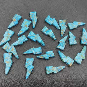 May include: A collection of turquoise-colored gemstone lightning bolt charms with gold-colored veining. The charms are arranged on a dark surface. These gemstone charms are ideal for jewelry making or craft projects.