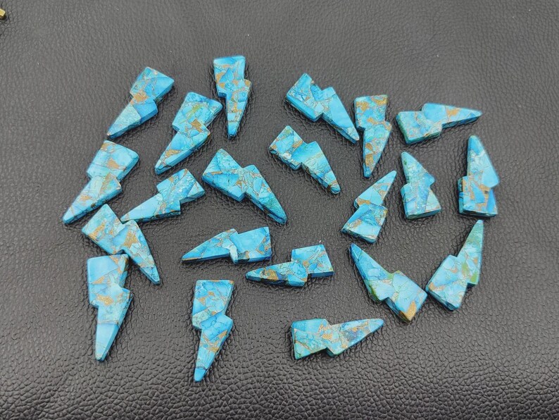 May include: A collection of turquoise-colored gemstone lightning bolt charms with gold-colored veining. The charms are arranged on a dark surface. These gemstone charms are ideal for jewelry making or craft projects.