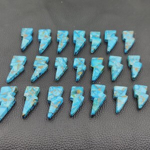 May include: Twenty-one turquoise-colored gemstone lightning bolt charms with gold-colored veining. The charms are arranged on a dark surface. Each charm is approximately 1 inch in length.