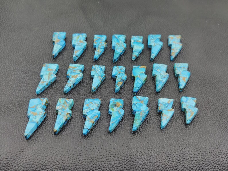 May include: Twenty-one turquoise-colored gemstone lightning bolt charms with gold-colored veining. The charms are arranged on a dark surface. Each charm is approximately 1 inch in length.