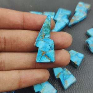May include: A collection of turquoise-colored gemstone pieces, each shaped like a lightning bolt. The stones have a vibrant blue hue with gold-colored veining throughout. The gemstones are displayed against a dark background.