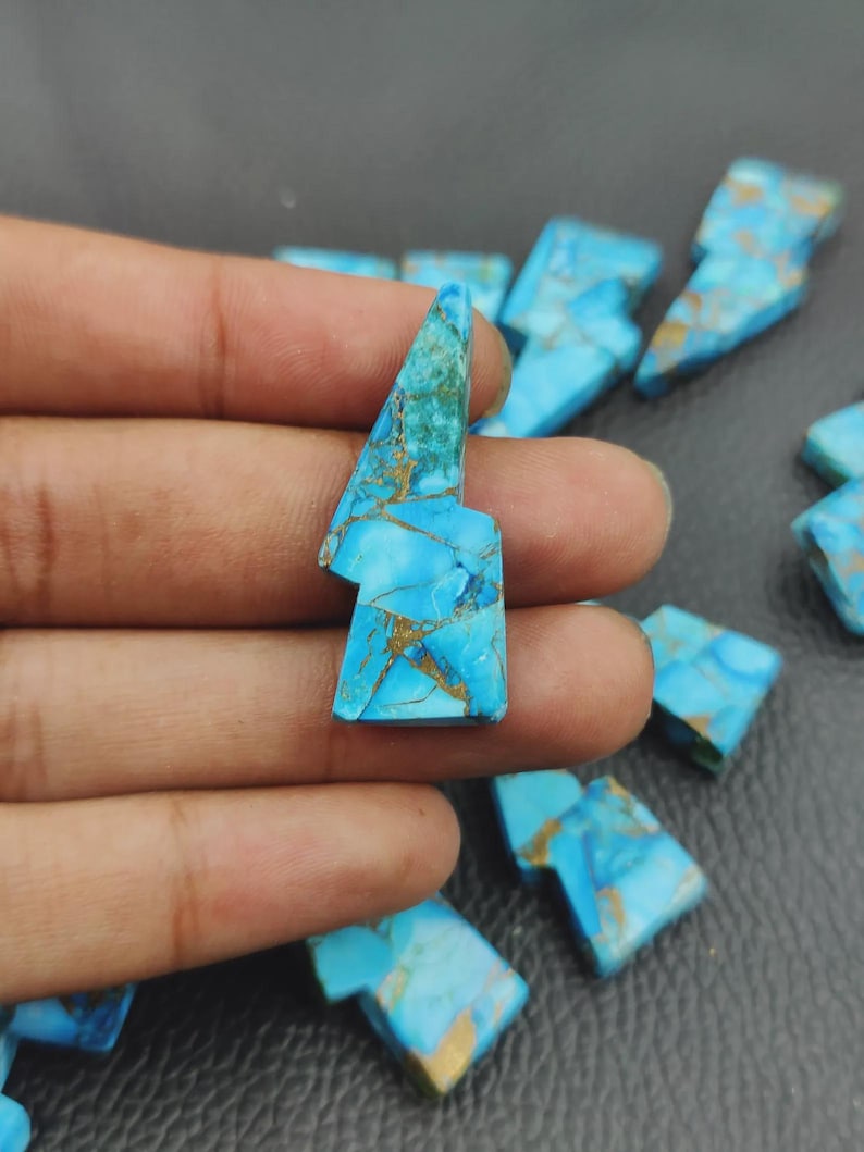 May include: A collection of turquoise-colored gemstone pieces, each shaped like a lightning bolt. The stones have a vibrant blue hue with gold-colored veining throughout. The gemstones are displayed against a dark background.