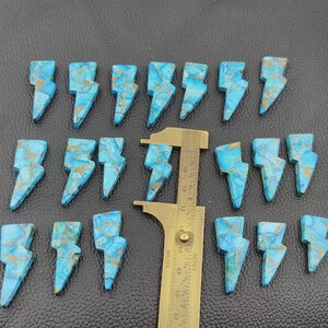 May include: A collection of turquoise-colored, lightning bolt-shaped gemstones with gold-colored veining. A brass ruler is placed among the stones. The gemstones are arranged on a dark, textured surface.