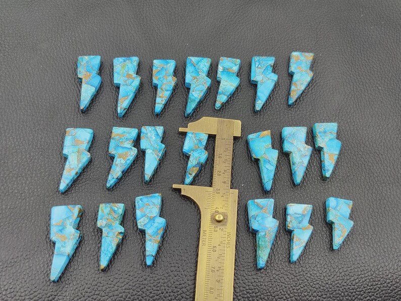 May include: A collection of turquoise-colored, lightning bolt-shaped gemstones with gold-colored veining. A brass ruler is placed among the stones. The gemstones are arranged on a dark, textured surface.
