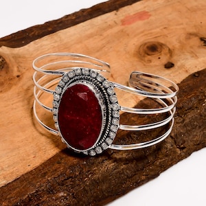 May include: Silver cuff bracelet with a large, oval, faceted, deep red gemstone. The gemstone is set in a detailed, floral-patterned bezel. The bracelet has multiple parallel silver bands.