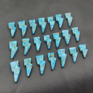 May include: Twenty-one turquoise and gold-colored gemstone lightning bolt charms arranged on a black leather surface. The charms are cut in a lightning bolt shape and have a marbled appearance.