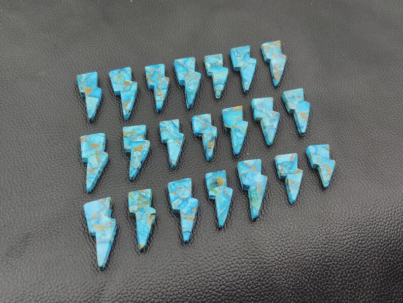 May include: Twenty-one turquoise and gold-colored gemstone lightning bolt charms arranged on a black leather surface. The charms are cut in a lightning bolt shape and have a marbled appearance.