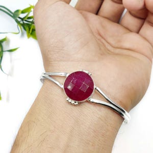 May include: A silver-toned cuff bracelet featuring a large, faceted, oval-shaped ruby gemstone. The ruby is a deep, rich red color. The bracelet has a simple, elegant design, with the gemstone as the focal point. The bracelet is worn on a wrist.