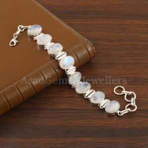 Pode incluir: Uma pulseira de prata com pedras preciosas de opala lunar ovais. A pulseira tem um design de corrente com opalas lunares engastadas em molduras de prata individuais. O fecho é do tipo lagosta. O texto "Asad Gem Jewellers" é visível.