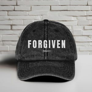 以下が含まれることがあります： 白い文字で「FORGIVEN」と書かれた黒い野球帽。 「FORGIVEN」の下には「PSALM 32：1」というテキストがあります。 白いレンガの壁の前に、白い表面に帽子が置かれています。