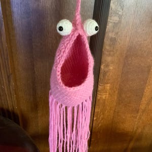 May include: A pink crocheted hanging with a large open mouth and two white googly eyes. The hanging has long pink fringe.