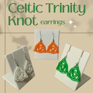 May include: Three pairs of Celtic Trinity Knot earrings in white, orange, and green, displayed on white stands. The earrings are triangular with a woven design. The text "Celtic Trinity Knot earrings" is at the top of the image.