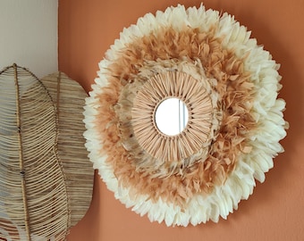 May include: Round wall mirror with a natural, bohemian design. The mirror is surrounded by a layered arrangement of white and brown feathers, with a woven raffia center. The mirror is mounted on a terracotta-colored wall.