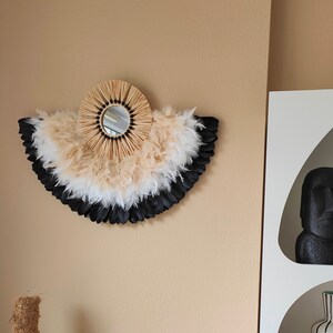 May include: A decorative wall mirror with a fan-shaped design. The mirror is surrounded by a circular frame of woven straw and a layered arrangement of black, white, and beige feathers. The mirror is mounted on a beige wall.